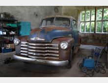 Ein alter, rostiger Pickup-Truck steht in einer Werkstatt mit offenem Fenster. Umgeben von Regalen, Werkzeugen und Maschinen, darunter ein Schweißgerät, liegt der Fokus auf dem Fahrzeug mit verblasster blau-brauner Lackierung.