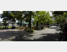 Ein gepflasterter Platz mit Bäumen, unter denen Tische und Stühle eines Straßencafés aufgestellt sind. Im Hintergrund befinden sich Gebäude und eine kleine, säulenartige Struktur, möglicherweise ein Pavillon.