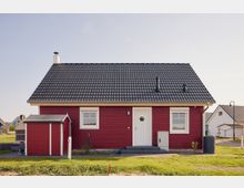 Einstöckiges rotes Einfamilienhaus mit Satteldach aus dunkelgrauen Ziegeln, weißem Eingang und zwei Fenstern an der Vorderseite. Links steht ein kleiner Anbau, umgeben von Rasen und Nachbarhäusern in einer ländlichen Wohngegend.