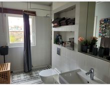 Modernes Badezimmer mit weiß gefliesten Wänden und gemustertem Fliesenboden. Ein Fenster lässt Tageslicht ein, daneben eine Duschkabine mit Vorhang, ein offenes Regal mit Handtüchern und Pflegeprodukten sowie ein Waschbecken mit Spiegel und Dekorationen.