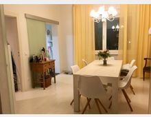 Esszimmer mit einem weißen Holztisch, umgeben von weißen Stühlen mit Holzbeinen. Im Hintergrund gelbe Vorhänge vor einem großen Fenster, ein Holztisch mit Bilderrahmen an der linken Wand und eine helle Hängelampe über dem Tisch.