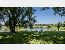 Grüne Parklandschaft mit Blick auf einen ruhigen See, umgeben von Schilf und Bäumen; im Vordergrund stehen mehrere Bänke und Tische sowie ein großer Baum, der Schatten spendet. Einige Menschen sitzen auf Stühlen und genießen die Aussicht.