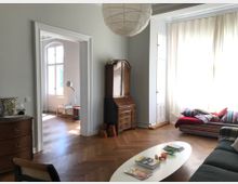 Wohnzimmer mit Fischgrät-Parkettboden, einer großen Fensterfront mit weißen Vorhängen, einem ovalen Couchtisch und einer mehrfarbigen Couch mit Kissen. Im Hintergrund ein angrenzender Raum mit Tageslicht, erkennbar durch eine breite Türöffnung. Ein antiker Holzschrank steht an der Wand rechts.