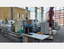 Ein Laborraum mit Industrieanlagen und technischen Geräten, darunter Rohrleitungen, Tanks und ein großer roter Trichter. Im Vordergrund stehen ein Schreibtisch mit Computer und ein Stuhl, während große Fenster und Ziegelwände den Raum umgeben.