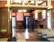 Ein historisch eingerichteter Raum mit verzierten Holzwänden, einer großen Doppeltür und roten Tapeten. Im Vordergrund stehen graue Stühle, und im Hintergrund hängen Gemälde sowie ein Kronleuchter von der Decke.