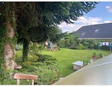 Garten mit Rasenfläche, großen Bäumen und dichtem Strauchwerk. Im Hintergrund ein Wohnhaus mit dunklem Schrägdach, eine Schaukel und eine Wäschespinne mit aufgehängter Kleidung.