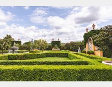 Gepflegter Barockgarten mit symmetrischen Hecken, einem zentralen Springbrunnen und dekorativen Orangeriebäumen. Im Hintergrund stehen mit Efeu bewachsene gelbe Wandelemente mit Statuen und Ornamenten.