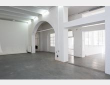 Heller, leerer Innenraum im Loft- oder Industrie-Stil mit weiß gestrichenen Wänden, Sichtdeckenbalken und grauem Betonboden. Große Rundbogenöffnung, breite Stützpfeiler und hohe Fenster lassen viel Tageslicht in den Raum.