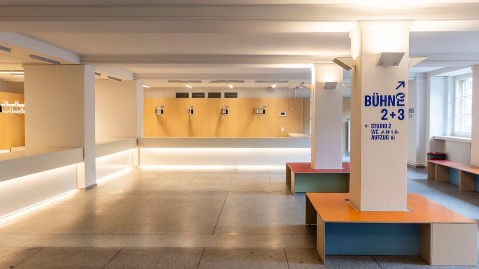 Heller, moderner Innenraum einer Veranstaltungs- oder Kulturstätte mit breitem Tresen vor einer Holzverkleidung, weiß gestrichenen Stützen und gefliestem Boden. Rechts stehen farbige Sitzbänke um eine Säule mit Wegweiser-Aufschrift „BÜHNE 2+3“, daneben große Fensterfront.