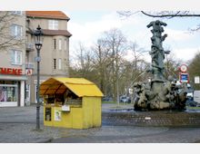 Ein kleiner Platz mit Kopfsteinpflaster, auf dem ein gelber Verkaufsstand für Honig steht. Daneben ein kunstvoll gestalteter Brunnen mit Figuren, im Hintergrund ein mehrstöckiges Gebäude mit rötlichem Dach und eine Apotheke.