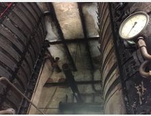 Industrieanlage mit einem großen, vertikalen Kessel, metallischen Rohren und Schläuchen. Auf der rechten Seite befindet sich ein rundes Manometer, und die Umgebung zeigt gealterte Wände und Industrial-Elemente.