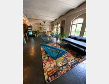 Industrieinspirierter Innenraum mit hohen Decken und großen Bogenfenstern, die viel Tageslicht hereinlassen. Im Vordergrund stehen bunte Möbel, darunter blaue Stühle und ein gemusterter Teppich; Pflanzen und Regale mit Dekorationen ergänzen die Einrichtung.