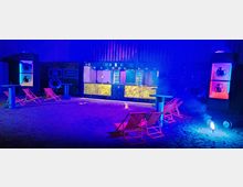 Ein Indoor-Bereich mit einer beleuchteten Bar im Hintergrund, umgeben von Liegestühlen und hohen Stehtischen; der Boden scheint sandig und die Szenerie wird von blauem und violettem Licht erhellt, ergänzt durch leuchtende Dekorationselemente.