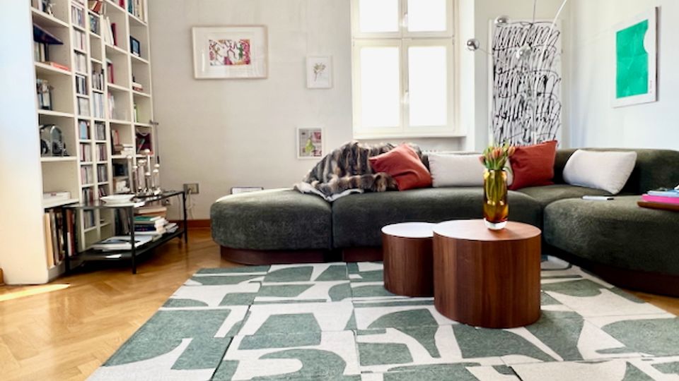 Helles, modernes Wohnzimmer mit großem L-förmigem Sofa, gemustertem Teppich und zwei runden Couchtischen aus Holz. Links stehen deckenhohe Bücherregale, hinten ein Fenster sowie Wandkunst und eine hohe Bogenlampe.