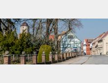 Straßenszene in einer Kleinstadt mit einem Fachwerkhaus im Vordergrund, umgeben von alten Bäumen. Im Hintergrund ist ein Kirchturm sichtbar, flankiert von weiteren Wohngebäuden in verschiedenen Pastellfarben.