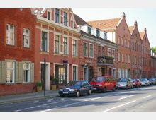 Eine historische Straßenszene mit Reihenhäusern aus rotem Backstein und Satteldächern, verziert mit weißen Fensterrahmen und Balkonen. Am Straßenrand stehen geparkte Autos entlang einer Fahrbahn mit markiertem Fahrradstreifen.