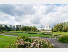 Weitläufiger Park mit gepflegten Grünflächen, Blumenbeeten und Wegen, im Hintergrund moderne Gebäude, darunter ein hoher Turm. Hohe Bäume säumen den Park, und der Himmel ist teils bewölkt.