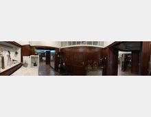 Innenansicht eines historischen Museums- oder Ausstellungsraums mit dunkler Holzvertäfelung, Parkettboden und Vitrinen sowie alten technischen Geräten und Messinstrumenten auf Tischen. Mehrere Türen und ein verglaster Durchgang verbinden den Raum mit angrenzenden Bereichen.