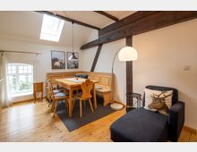 Helles Wohnzimmer mit einer Essecke aus hellem Holz, bestehend aus einer Eckbank, einem Tisch und Stühlen, auf Holzdielenboden. Sichtbare Holzbalken an der Decke, ein großes Oberlicht sowie ein Sessel mit Hirschkissen und Stehlampe ergänzen die Einrichtung, daneben ein rechteckiges Fenster mit Blick auf einen Garten.