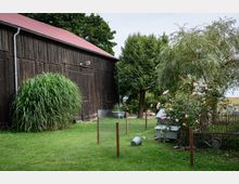 Hofbereich mit einer hölzernen Scheune mit rotem Dach auf der linken Seite, umgeben von Grünflächen, Bäumen und Sträuchern. Im Vordergrund befinden sich ein Maschendrahtzaun, ein Eimer, blühende Rosensträucher und ein kleiner hölzerner Unterschlupf.