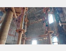 Innenansicht eines Hochseilgartens in einem historischen Backsteingebäude mit hohen Decken. Zu sehen sind Holzkonstruktionen, Kletterelemente wie Netze und Seile sowie eine Wendeltreppe, durch die Sonnenlicht durch große, bogenförmige Fenster fällt.