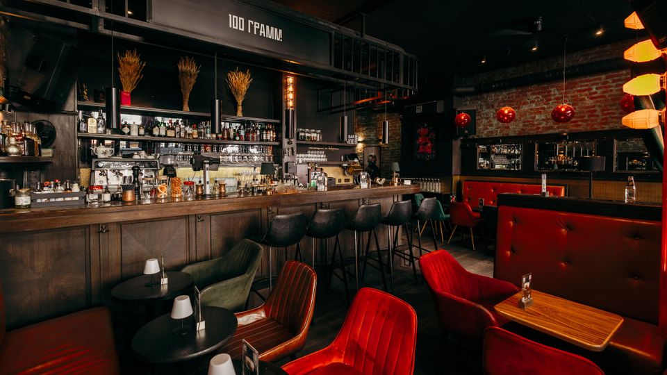 Innenraum einer Bar im Industriestil mit langer Holztheke, hohen Barhockern und Regalen voller Flaschen vor schwarzer Wand mit Beschriftung „100 GRAMM!“. Im Sitzbereich stehen rote Polsterbänke und rote Sessel an kleinen Tischen, ergänzt durch Backsteinwand, dunkle Decke und warme Hängeleuchten.
