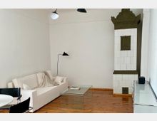 Wohnzimmer mit minimalistischem Design, bestehend aus einem weißen Sofa, einem Glastisch und einer Stehleuchte. Rechts befindet sich ein historischer, verzierten Kachelofen, der mit weißen und olivgrünen Fliesen verkleidet ist, auf einem Boden aus Holzplanken.