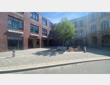 Ein gepflasterter Hof mit einem Baum in der Mitte, umgeben von Gebäuden im Backstein- und klassizistischen Stil. Die Backsteinfassade zeigt Schilder wie „Tischlerei Wiedmann“ und „Yoga“, während vor dem anderen Gebäude Sitzmöglichkeiten und ein Kunstobjekt zu sehen sind.