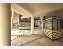 Weitläufiger Innenraum eines Gebäudes mit einer Kombination aus moderner und historischer Architektur. Im Vordergrund befinden sich runde Informations- und Kassenschalter, dahinter sind Ziegelwände mit hohen Fenstern zu sehen.