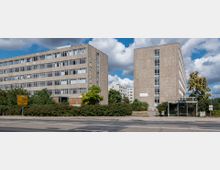 Zwei mehrstöckige, rechteckige Gebäude mit einer grauen, grobkörnigen Fassade und vielen schmalen Fenstern, umgeben von Büschen und Bäumen. Im Vordergrund befindet sich eine Straße mit einem Gehweg und einer Bushaltestelle mit Glasüberdachung.