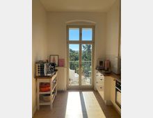 Bright, narrow kitchen with white cabinets, wooden countertops, and a small side cart, centered on tall French doors with an arched transom window. The doors open to a small balcony with railing and seating, overlooking leafy trees and nearby buildings.