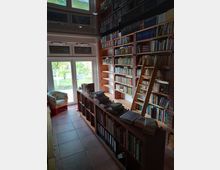 Eine kleine Bibliothek mit hohen Bücherregalen aus Holz, die bis zur Decke reichen, und einer schmalen Leiter. Im Vordergrund steht ein niedriger Holztresen mit Büchern und Ordnern, im Hintergrund sind Glasfenstertüren mit Blick ins Grüne sowie ein Sessel sichtbar.