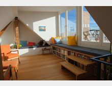 Wohnzimmer in einer Dachgeschosswohnung mit Holzboden, einem großen Fenster mit Blick auf eine Terrasse und einer Fensterbank, die als Sitzgelegenheit mit bunten Kissen gestaltet ist. Darunter befinden sich Regale mit Büchern, daneben ein kleiner Holztisch, ein Sessel und ein schräger Holzbalken als architektonisches Detail.