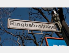 Straßenschild mit der Aufschrift „Ringbahnstraße“, montiert an einem Laternenmast vor einem blauen Himmel mit kahlen Ästen im Hintergrund.