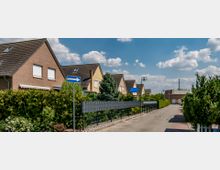 Reihenhäuser in einer Wohnsiedlung mit Ziegel- und Putzfassaden, dunklen Schrägdächern und Vorgärten, die durch Hecken und Zäune abgetrennt sind; eine gepflasterte Einbahnstraße führt durch die Szene, umgeben von Bäumen und Büschen, bei klarem Himmel.