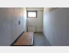 Schmaler, karger Raum mit rau verputzten weißen Wänden, einem kleinen vergitterten Fenster an der Stirnwand und einem Metallbett mit Holzlatten auf dem grauen Betonboden. Unter dem Fenster befindet sich ein Heizkörper mit freiliegenden Rohren.