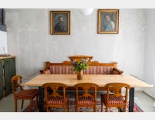 Holzbank mit rot gestreifter Polsterung, flankiert von vier passenden Holzstühlen, an einem schlichten Holztisch in einem Raum mit neutraler Wandfarbe; darüber zwei gerahmte Porträts an der Wand und ein Blumenstrauß in der Tischmitte. Links im Hintergrund ein kleines Spülbecken mit grünem Unterschrank.