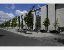 Moderne Büro- oder Regierungsgebäude mit einer Fassade aus Beton und großen Glasflächen. Davor verläuft eine breite Straße mit parkenden Autos, angelegten Bäumen, Fahrradständern und Gehwegen.