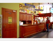 Rezeption in einer Bowlinghalle mit einem langen Tresen aus Holz, mehreren Schildern wie „Schuhausgabe“ und „Anmeldung/Kasse“ sowie dekorativen Elementen und Fotos an der gelb gestrichenen Wand. Ein Mann steht vor dem Tresen, daneben eine Tür mit der Aufschrift „Privat“.