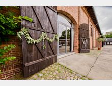 Backsteinbau mit großen Glastüren und dekorierten Holztoren, geschmückt mit Blumen- und Blättergirlanden. Der Boden besteht aus Kopfsteinpflaster und Beton, umgeben von Grünflächen und Topfpflanzen.