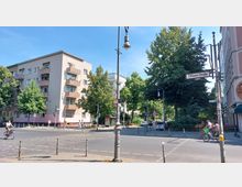 Straßenkreuzung in einer städtischen Umgebung mit einem vierstöckigen Wohngebäude auf der linken Seite, mehreren Bäumen und einem Straßenschild mit der Aufschrift „Ohlauer Straße“. Im Vordergrund befinden sich Fahrradfahrer, Fußgänger und Laternenmasten.