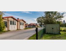 Straßenszene mit alten Backsteingebäuden und Wellblechtoren auf der linken Seite, daneben ein Zaun und ein Baum. Im Hintergrund sind kleinere Gebäude mit roten Dächern, ein Wasserturm und Wiese zu sehen.