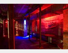 Unterirdischer Raum mit roher Beton- und Ziegelarchitektur, beleuchtet von roten und blauen Lichtern. Die Einrichtung umfasst Metallstützen, schmale Tische und Bänke, mit einem industriellen Ambiente.