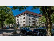 Mehrstöckiges Wohngebäude mit heller Fassade und roten Ziegeldach, umgeben von Bäumen und parkenden Autos an einer mit Kopfsteinpflaster versehenen Straße. Schilder und Laternen im Straßenbereich vervollständigen die städtische Umgebung.