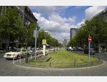 Straßenansicht einer breiten Allee mit mittlerem Grünstreifen, umgeben von Gebäuden und Bäumen. Im Hintergrund ist eine markante Kirchturmruine sichtbar, während Autos und Verkehrsschilder die Szene prägen.