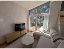 Heller Wohnbereich in einem Holzhaus mit weiß vertäfelten Wänden und schräger Decke, Sofa, Couchtisch und TV auf Lowboard. Große bodentiefe Fensterfront mit Oberlicht und weißen Vorhängen führt auf eine Holzterrasse mit Blick auf Bäume.