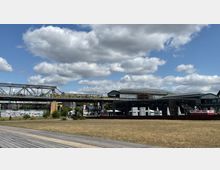 Weitläufige Szene mit Wiese und Holzstegen im Vordergrund, darüber eine Hochbahnbrücke mit gelbem Zug. Im Hintergrund stehen Industriebauten und moderne Gebäude, teils mit Glas- und Stahlkonstruktionen, bei leicht bewölktem Himmel.