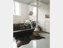 Modernes Wohnzimmer mit minimalistischer Einrichtung. Zu sehen sind ein dunkelgraues Sofa mit Kissen, ein Teppich in Felloptik, eine Hängelampe, Würfelmöbel und Zimmerpflanzen vor weißen Wänden und großen Fenstern.