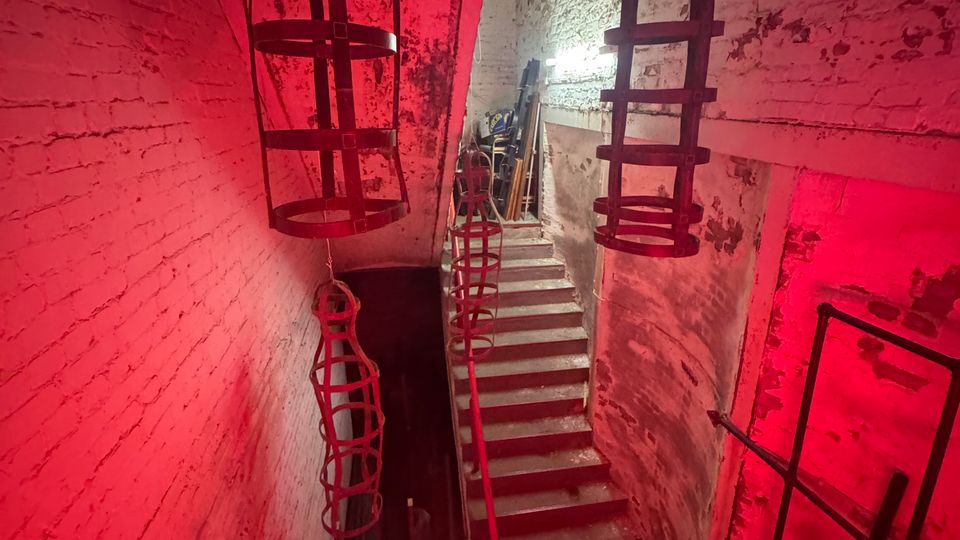 Ein schmaler, industriell wirkender Treppenaufgang in einem Gebäude mit weißen, teils verwitterten Ziegelwänden, beleuchtet von roten Lichtern. Metallstrukturen hängen von der Decke, und der Raum wirkt wie ein Lager- oder Werkstattbereich.