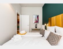 Modernes Schlafzimmer mit Doppelbett, weißer Bettwäsche und senfgelbem, gepolstertem Kopfteil vor einer dunkelgrünen Akzentwand. Neben dem Bett stehen eine kleine Wandlampe und ein Heizkörper; gegenüber sind eine offene Tür, ein schmaler Beistelltisch mit Skulptur sowie ein gerahmtes Bild an der Wand zu sehen.